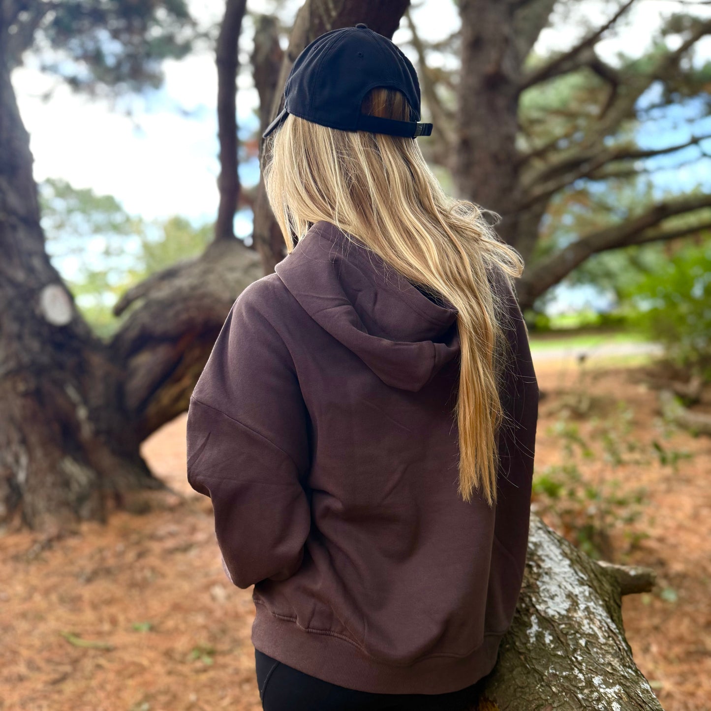 Person wearing a hoodie and black cap leaning against a tree in a forest setting