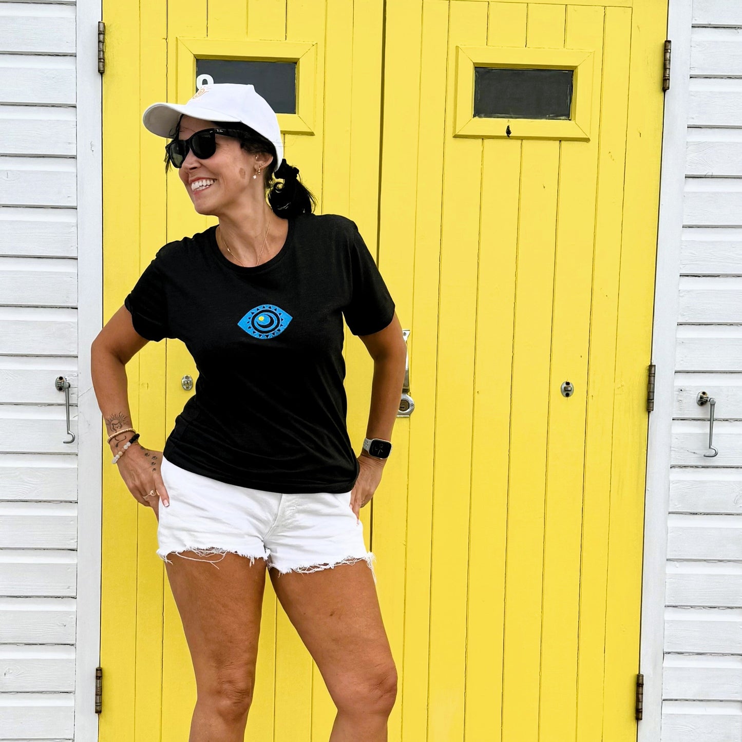 Woman wearing a black t-shirt with a blue logo and white shorts standing in front of a yellow door.