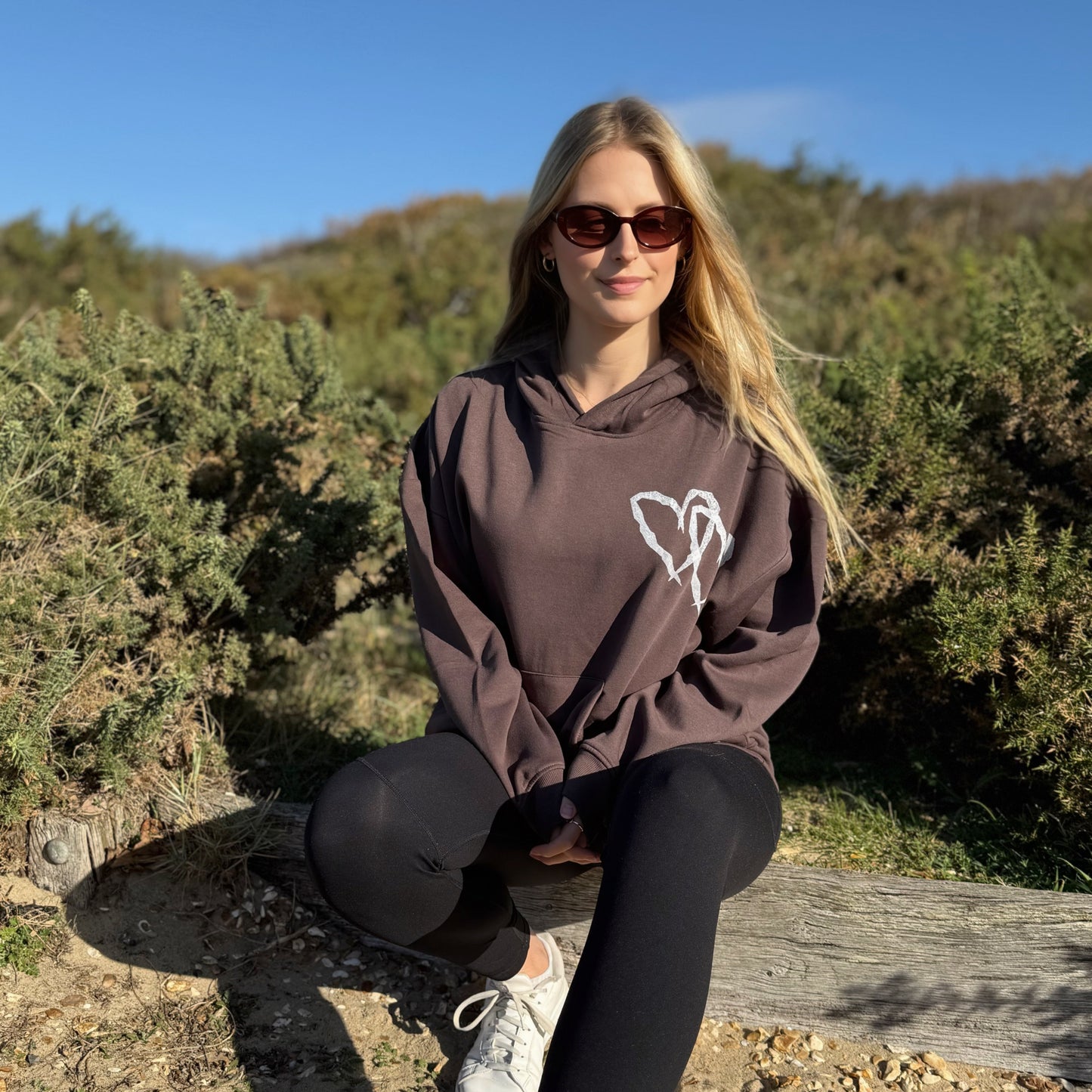 Woman sitting outdoors wearing a brown hoodie with a white logo, sunglasses, and black leggings.