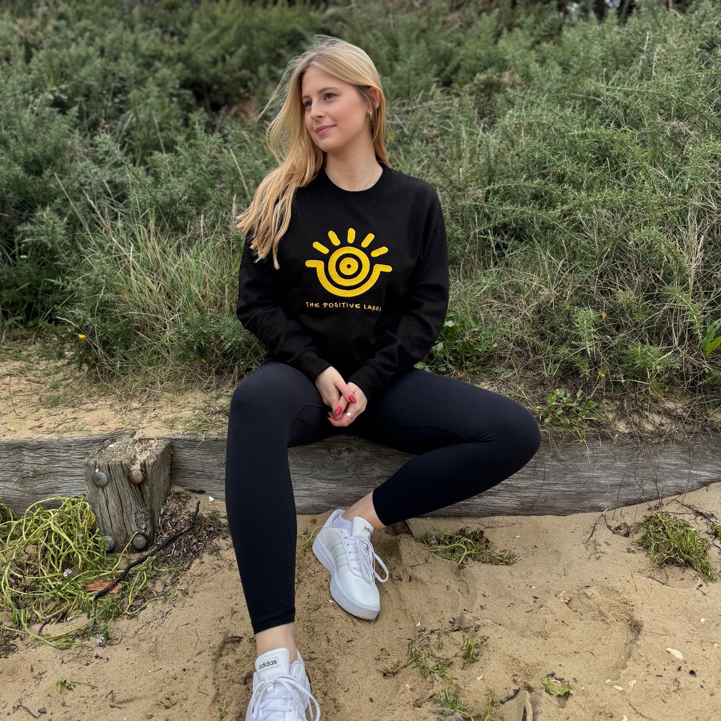 A woman sitting on a wooden bench in a natural outdoor setting, wearing a black sweatshirt with a gold sun emblem and the text 'The Positive Label', paired with black pants and white sneakers.