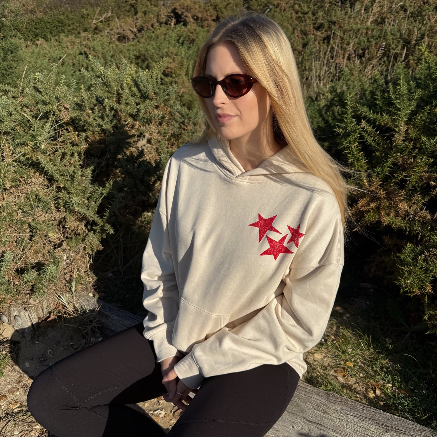 Woman wearing a beige hoodie with red star designs, sitting outdoors.