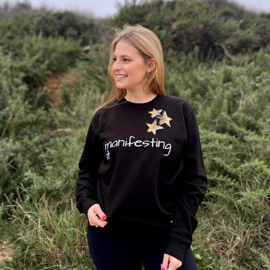 A woman standing in a natural outdoor setting, smiling while wearing a black sweatshirt with the text 'MANIFESTING' and a star design on the front.