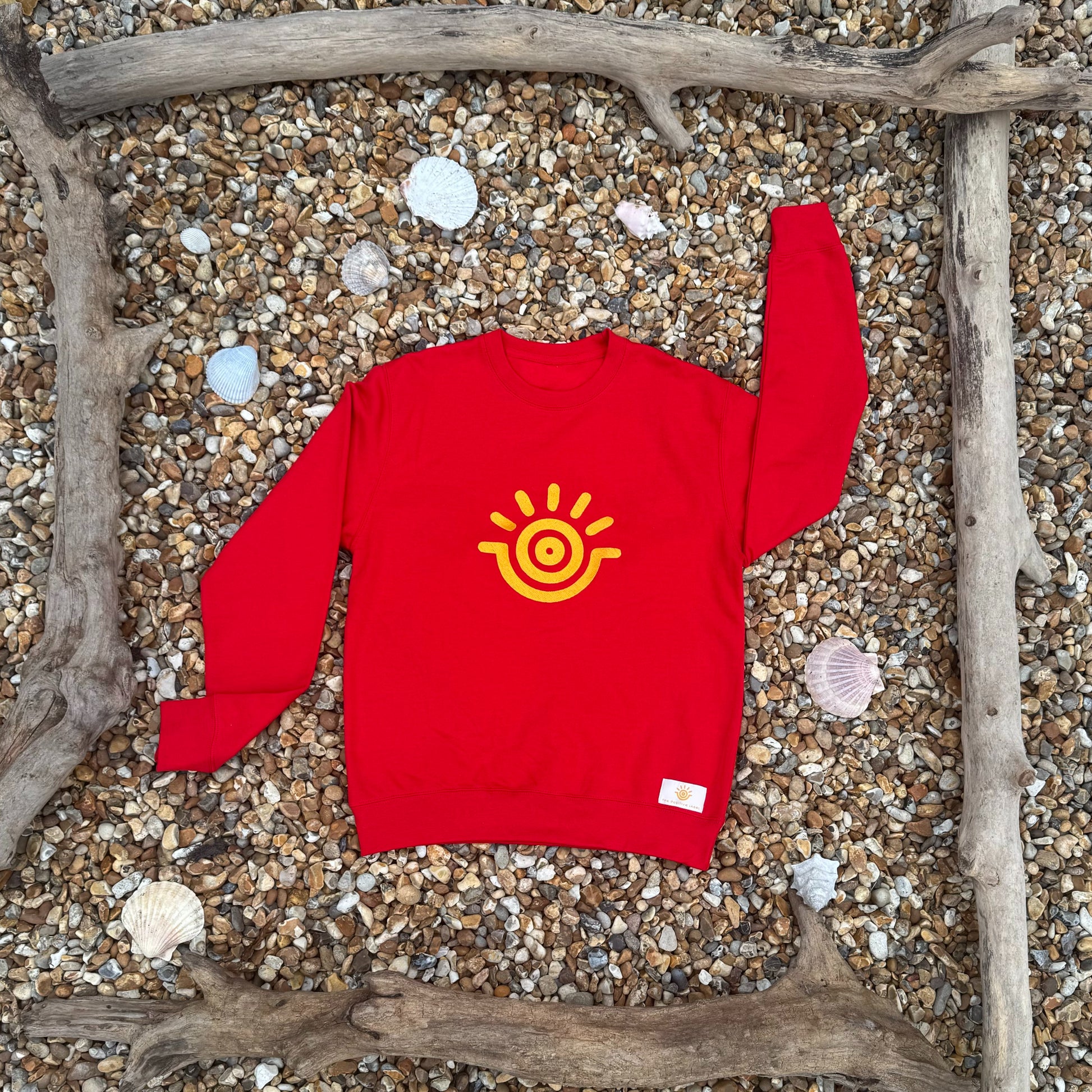 A red sweatshirt with a golden yellow sun emblem in gold embroidery, displayed on a wooden frame with seashells and driftwood in the background.