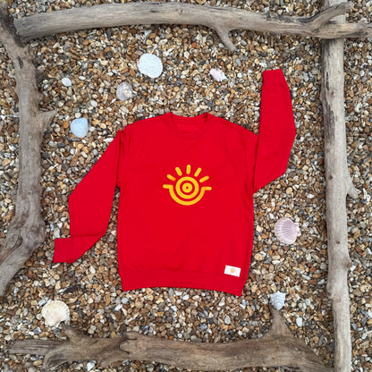 A red sweatshirt with a golden yellow sun emblem in gold embroidery, displayed on a wooden frame with seashells and driftwood in the background.