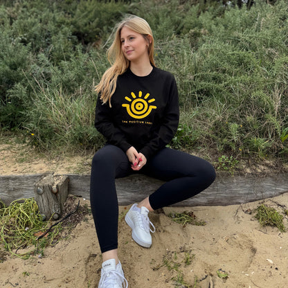A woman sitting on a wooden bench in a natural outdoor setting, wearing a black sweatshirt with a gold sun emblem and the text 'The Positive Label', paired with black pants and white sneakers.