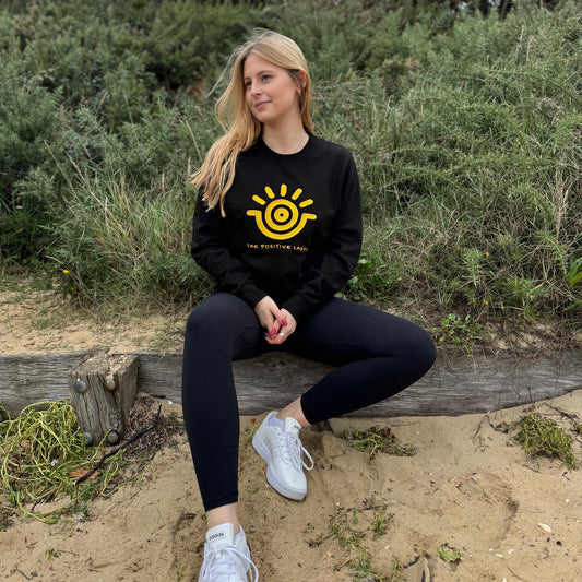 A woman sitting on a wooden bench in a natural outdoor setting, wearing a black sweatshirt with a gold sun emblem and the text 'The Positive Label', paired with black pants and white sneakers.