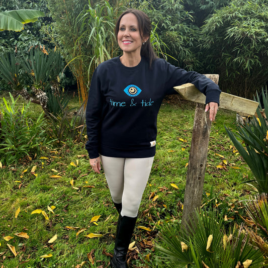 A woman standing by a wooden post in a garden-like setting, wearing a navy sweatshirt with turquoise embroidered text and a hand-painted eye symbol, paired with light-colored riding pants.