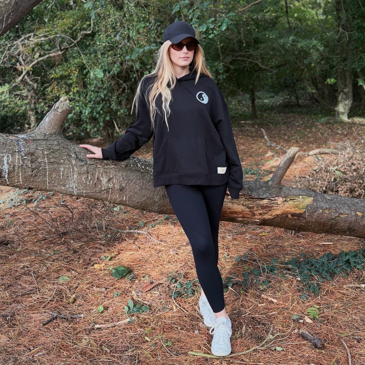 Person wearing a black hoodie and leggings standing next to a fallen tree in a forest.
