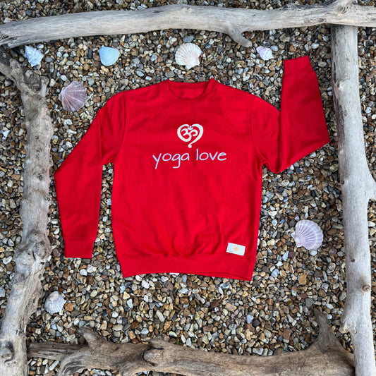 A red sweatshirt with the text 'YOGA LOVE' and a heart symbol embroidered on the front, displayed on a rocky surface with seashells and a wooden branch in the background.