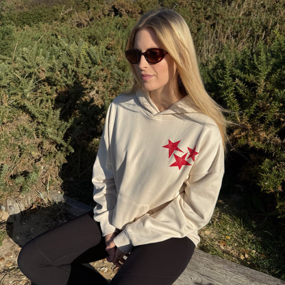Woman wearing a beige hoodie with red star designs, sitting outdoors.